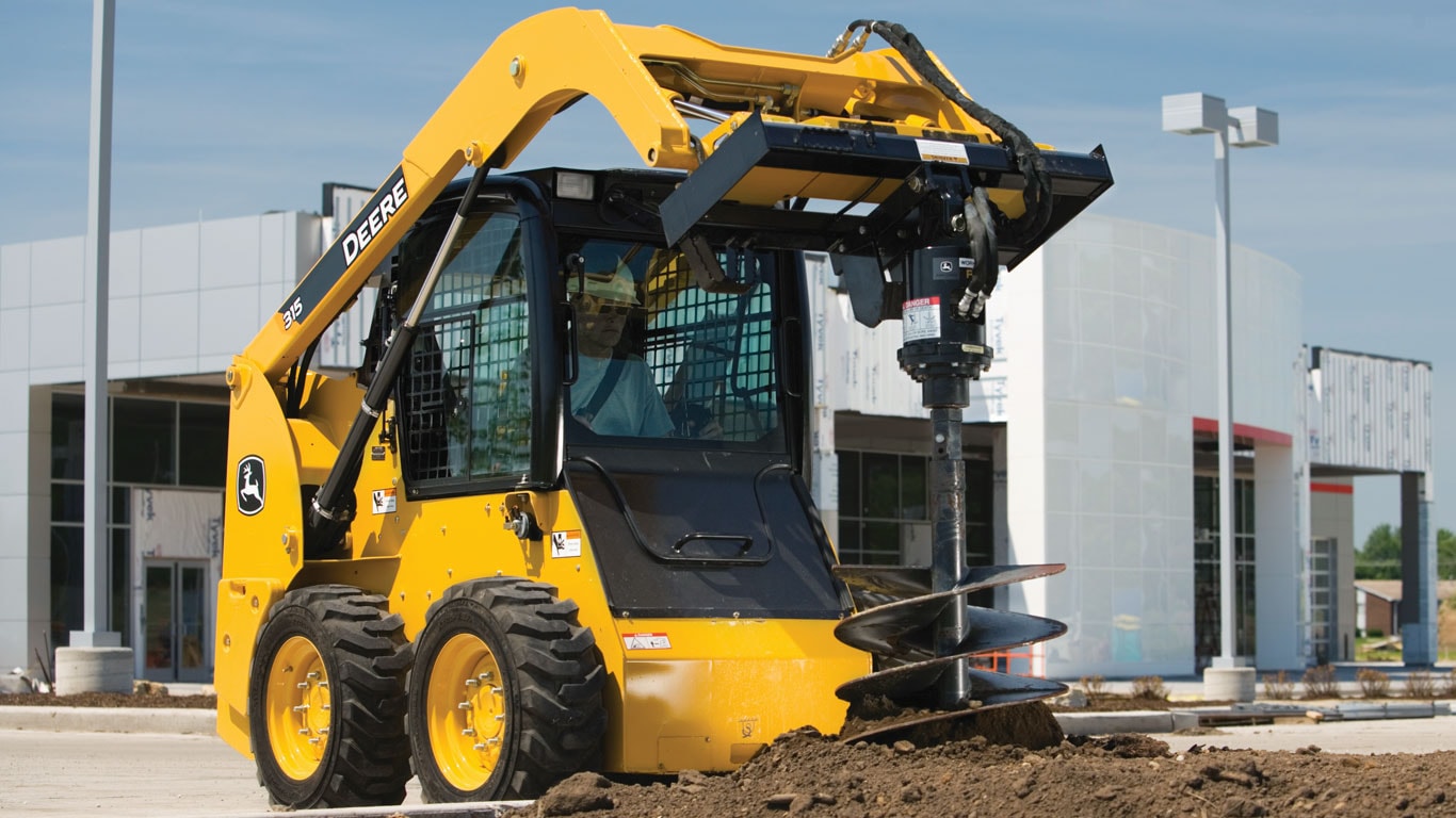 Une chargeuse à direction différentielle John Deere équipée de l’accessoire tarière à transmission à engrenage planétaire creuse sur un chantier.